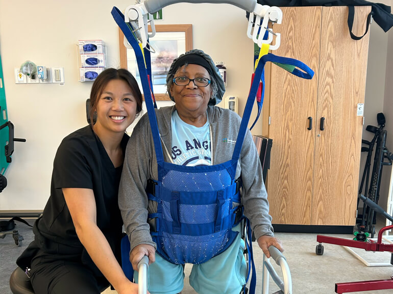 Carol Roseboro uses a supportive device to relearn to walk after amputation with her therapist by her side.