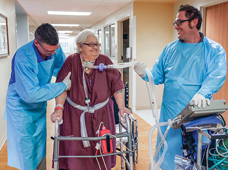 Patient on a vent walks down the hallway with help from therapists.