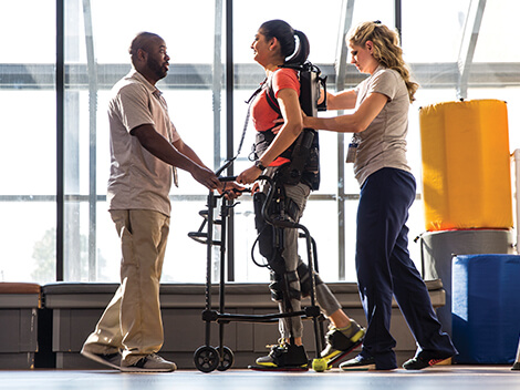 Patient wears robotics while therapists help her walk. 