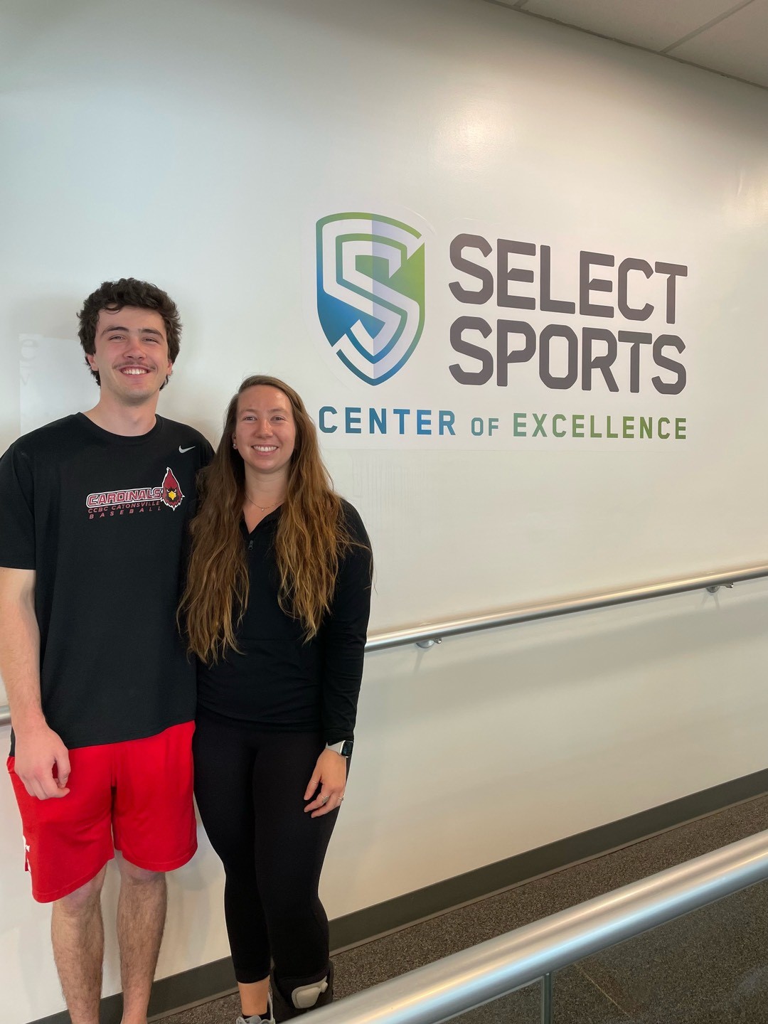 Liam, a patient, posing for a photo with his physical therapist in front of the Select Sports logo. 