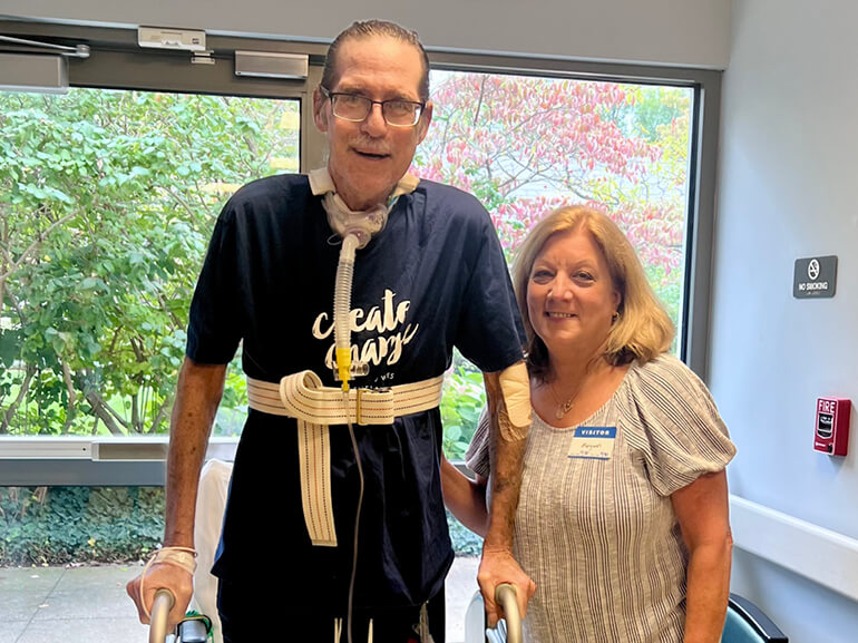 Carl stands with is wife in the hospital hallway using a walker for support.