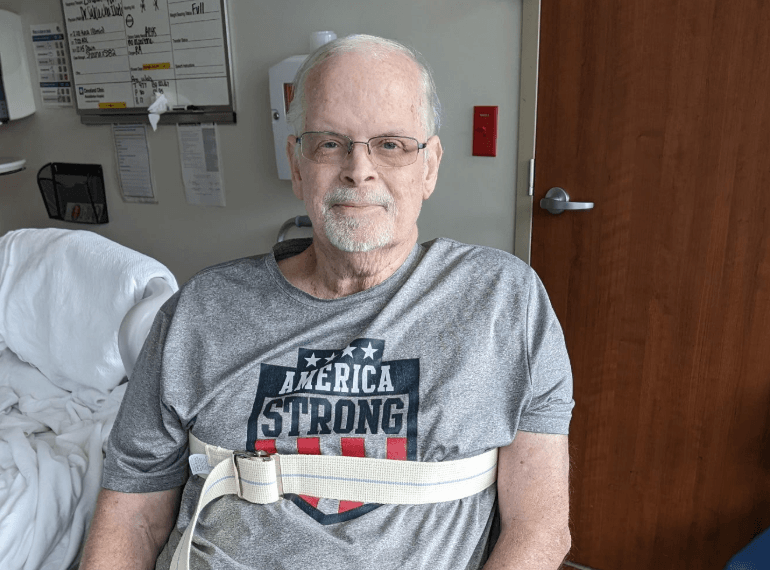 Glenn George sits in a hospital room, wearing a weight belt and a T-shirt and glasses. 