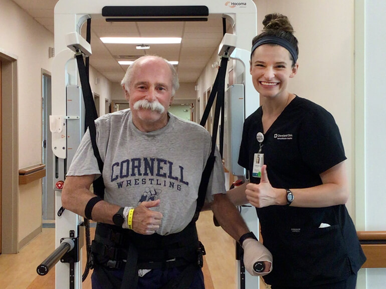 Jere stands utilizing weight supporting equipment while giving a thumbs up to the camera along with his therapist.