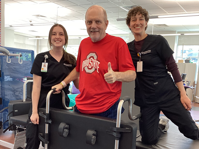 Larry stands using his walker for support with two of his therapists. He's giving the camera a thumbs up.