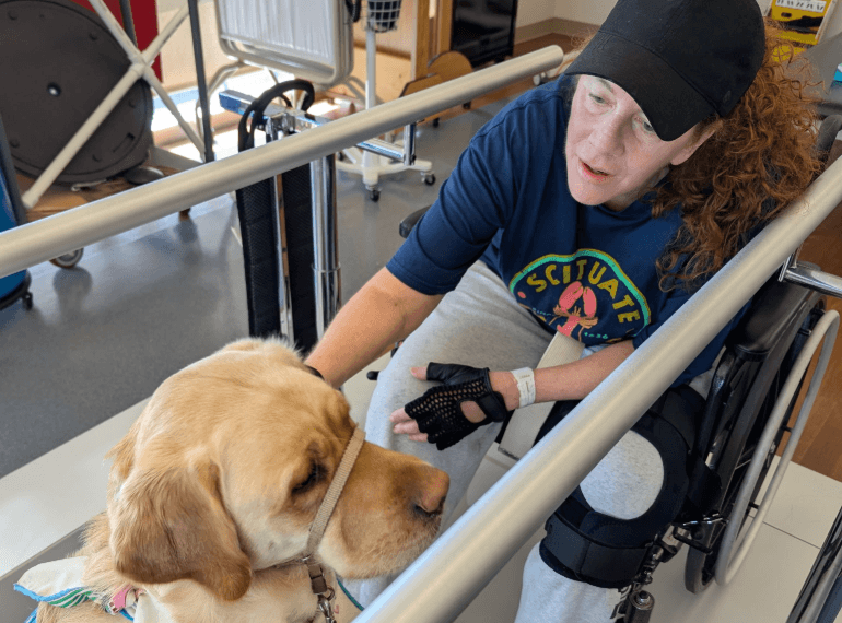Mary Apicella leans forward between parallel bars and pets a therapy dog. Mary wears a baseball cap. 