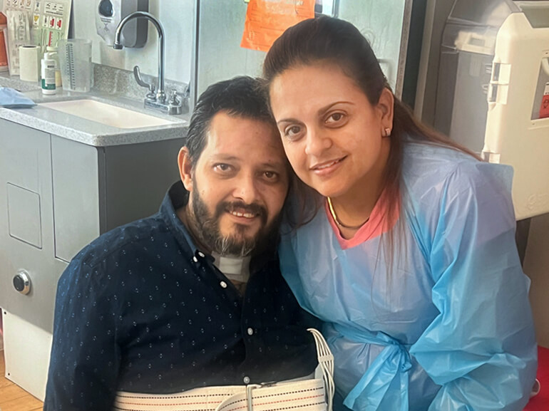 Miguel sits in a wheel chair in his hospital room posing with his wife to smile at the camera.