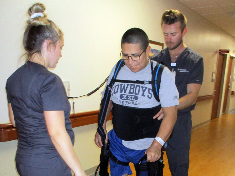 Raul uses an assistive device and the help of his care team to walk down the hospital hallway.