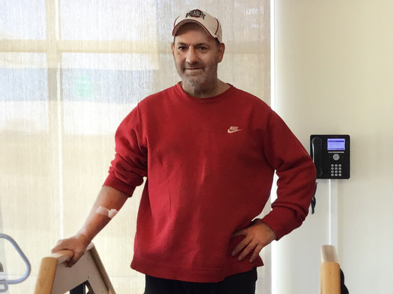 Richard Vojtush stands in a therapy gym in a red sweatshirt with his hand on his hip.