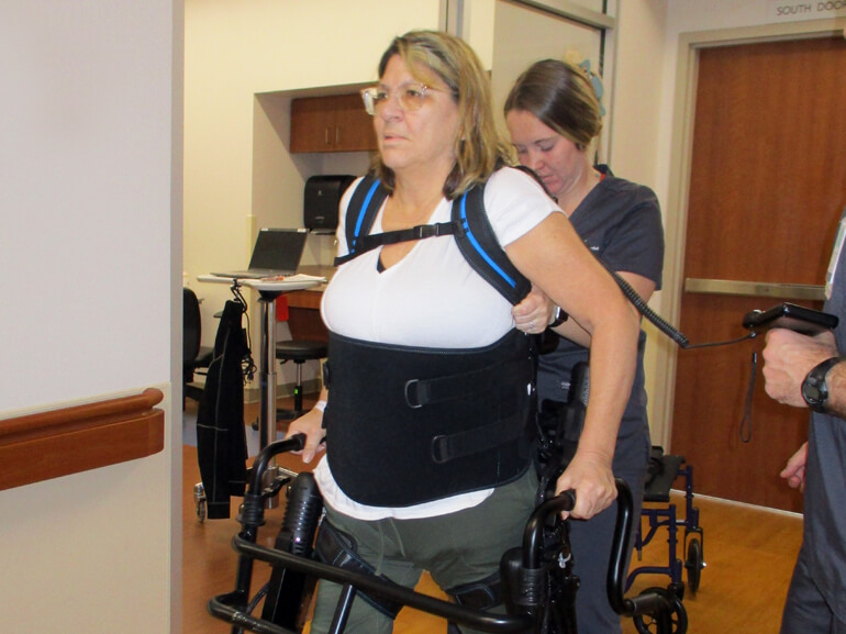 Sandra uses a walker and support brace to walk down a hospital hallway.