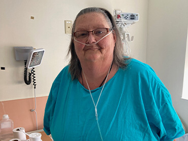 Lynn smiling and sitting in a hospital room, wearing glasses and a teal shirt.