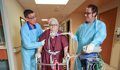 Women with a trach walks down a hallway using a cane with her care team.
