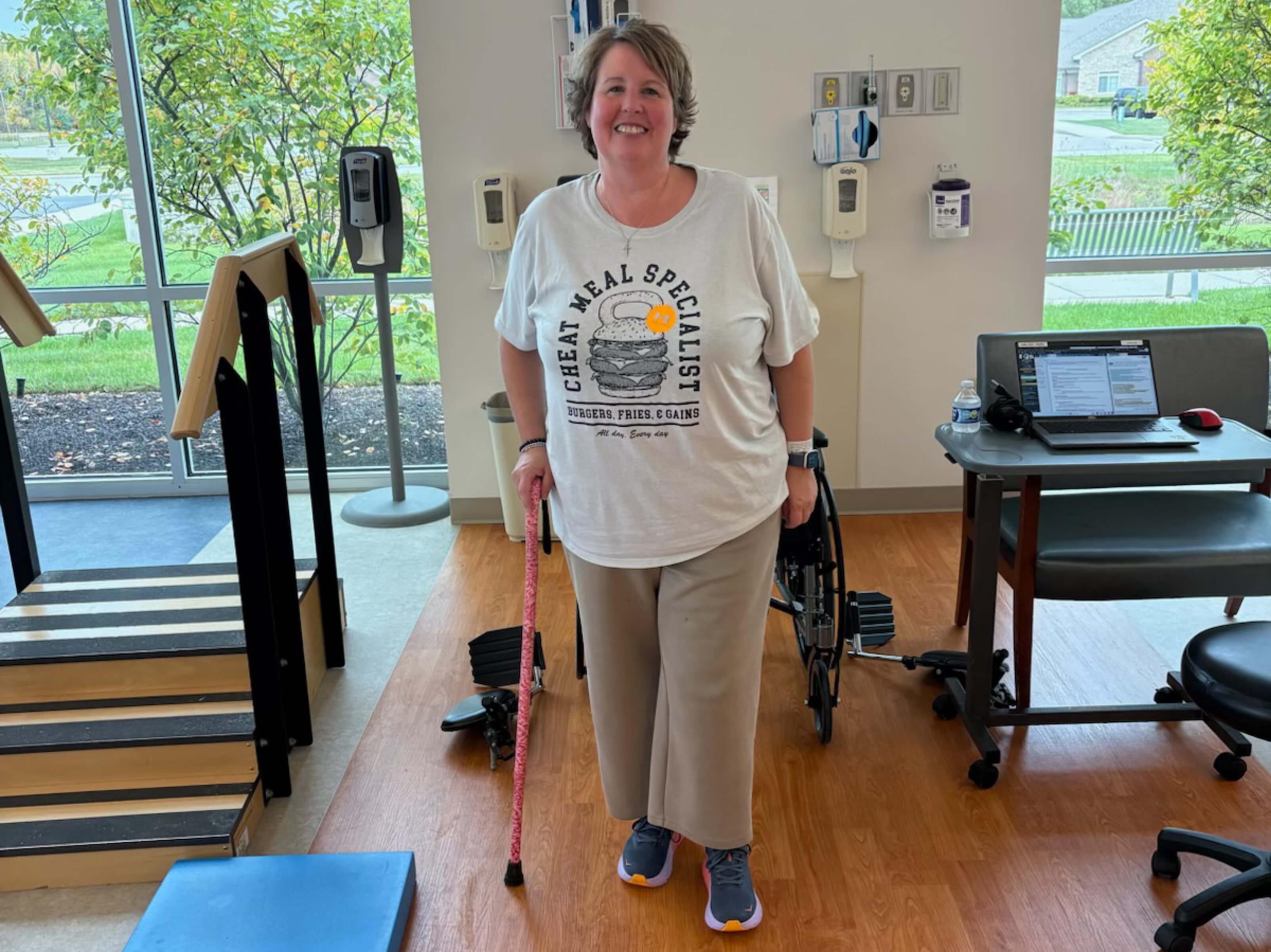 Angie Fairbanks smiles as she steps forward. She is wearing sweat pants and a T-shirt in a therapy gym, and she walks with the support of a cane. Behind her, two windows look out on green trees.