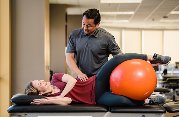 A woman is laying on her side on a table with a yoga ball in between her legs. Her therapist is standing beside