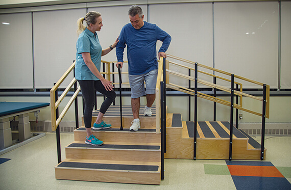 A man is walking down a set of stairs with a cane as a therapist stand beside for support