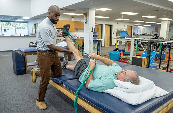A man is laying on his back on a table and holding a stretch band that is hooked onto his foot. A therapist is standing beside and holding the patient's leg in the air