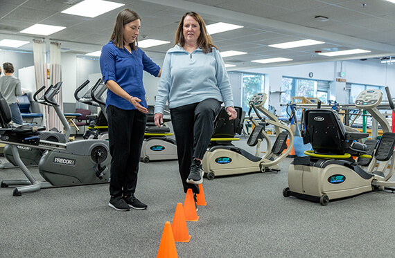 A woman is standing in front of multiple cones and standing on one leg while the elevated foot taps the cone. A therapist is standing beside for support