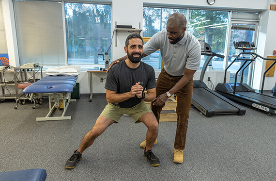 A man is in a side lunge inside of a gym as a therapist is standing by for support