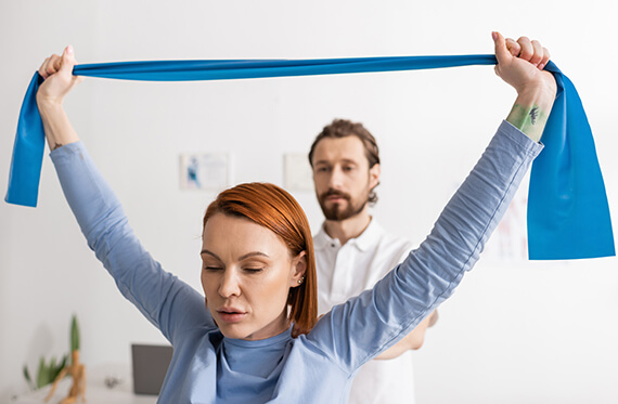 Close up image of woman pulling therapy band over her head while her therapists watches. 