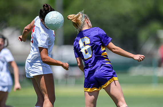 Two girl soccer players are on the field trying to hit a soccer ball with their heads