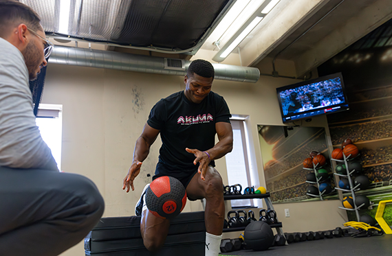 An athlete is performing split squat while throwing a medicine ball on the ground. A physical therapist is sitting beside him slightly off frame