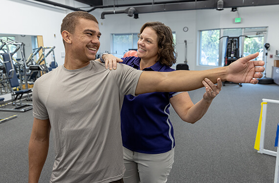 A man is lifting one arm straight out to his side with a physical therapist standing behind him