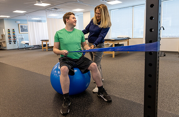 A man is sitting on a medicine ball and pulling on a resistance band while a physical therapist stands beside him