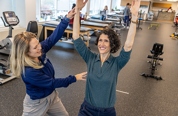 A woman is standing and lifting both arms overhead while a physical therapist is standing beside