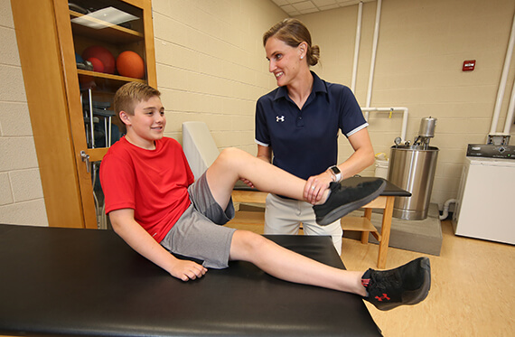 A young man is sitting on a table while a physical therapist lifts one of his legs up 