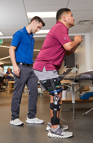 A man has a knee brace on and performing standing exercises with a physical therapist beside him