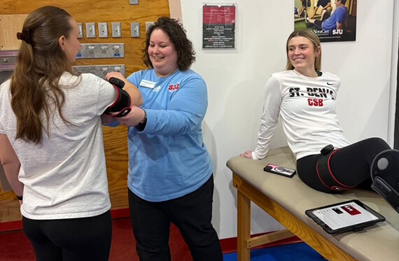 Corinne is standing beside an athlete and reading the athlete's blood pressure while another athlete sits on a table beside them