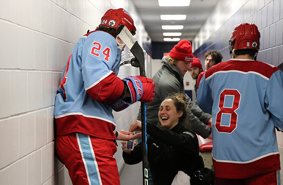 Holly is kneeling on the ground and surrounded by hockey players. She is smiling as she hands a hockey player medicine