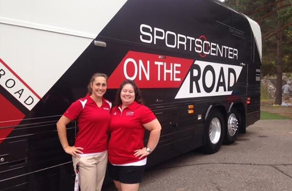 Holly and Corinne stand beside each other in front of a travel bus and smile at the camera