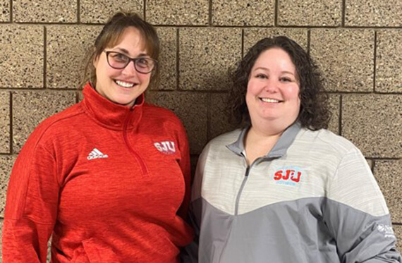 Holly and Corinne stand beside each other in front of a brick wall and smile at the camera