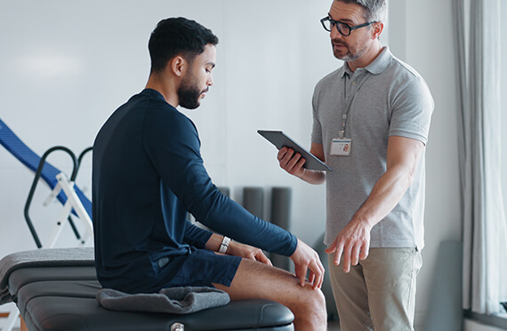 A therapist has an iPad in hand and is speaking to a patient that is sitting on an exam table while pointing to his knee