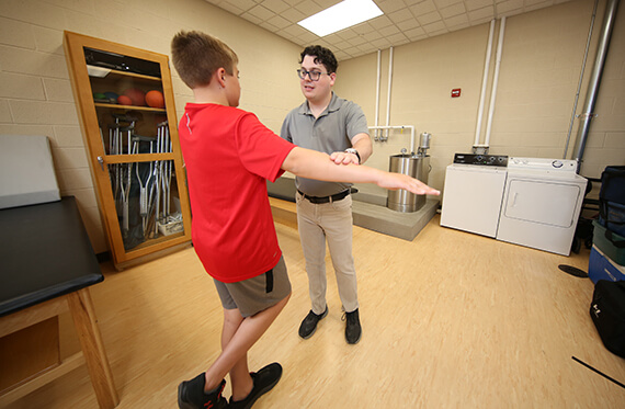 A boy is standing on one leg with both arms out to his side. A therapist is standing in front of him