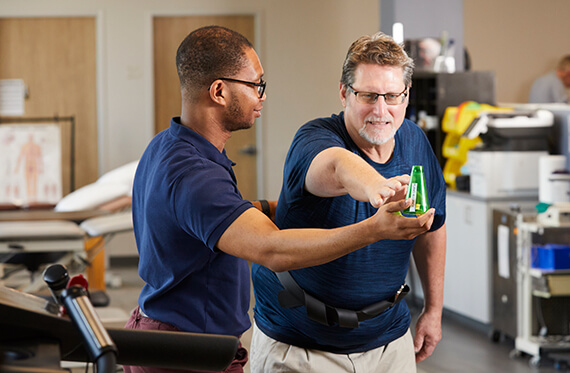 A therapist is holding out a green object in front of a patient that is using on arm to reach forward at the green object