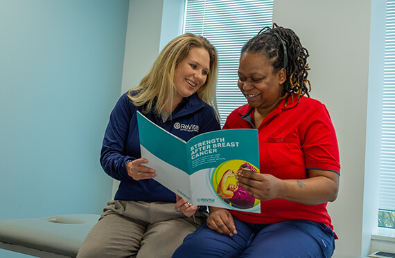 A therapist and patient are sitting beside each other on a table and reading through a cancer rehabilitation pamphlet