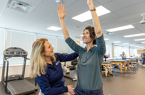 A therapist is holding her hands out to support a patient that is standing up and reaching for the sky with both hands