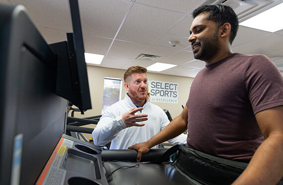 A close up of a physical therapist standing next to a patient as the patient is walking on a treadmill