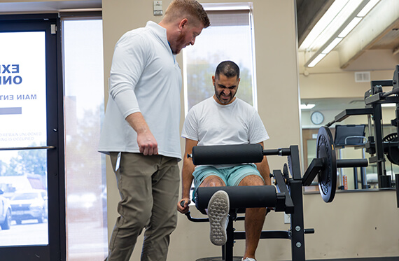 A physical therapist is standing beside a patient as the patient is completing a leg extension on a machine