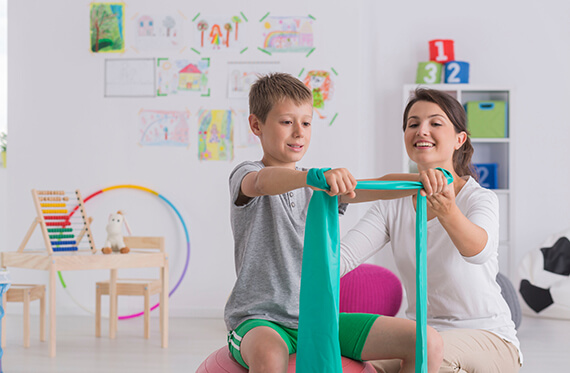 A young boy using bands to exercise his upper body while a therapist is beside him