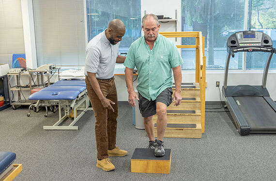 A patient has one foot on a block and about to step up onto the block. A therapist is standing beside for support