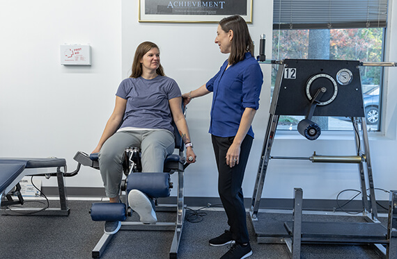A woman is raising one leg on a leg extension machine while a therapist is standing beside for support