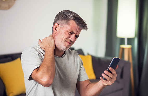 A man is sitting on a couch and holding one hand on his neck in pain while looking at his phone