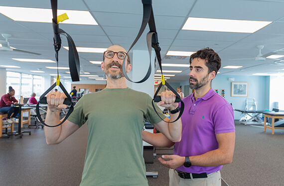 a patient is using TRX bands to completed pullups. A therapist is standing beside for support